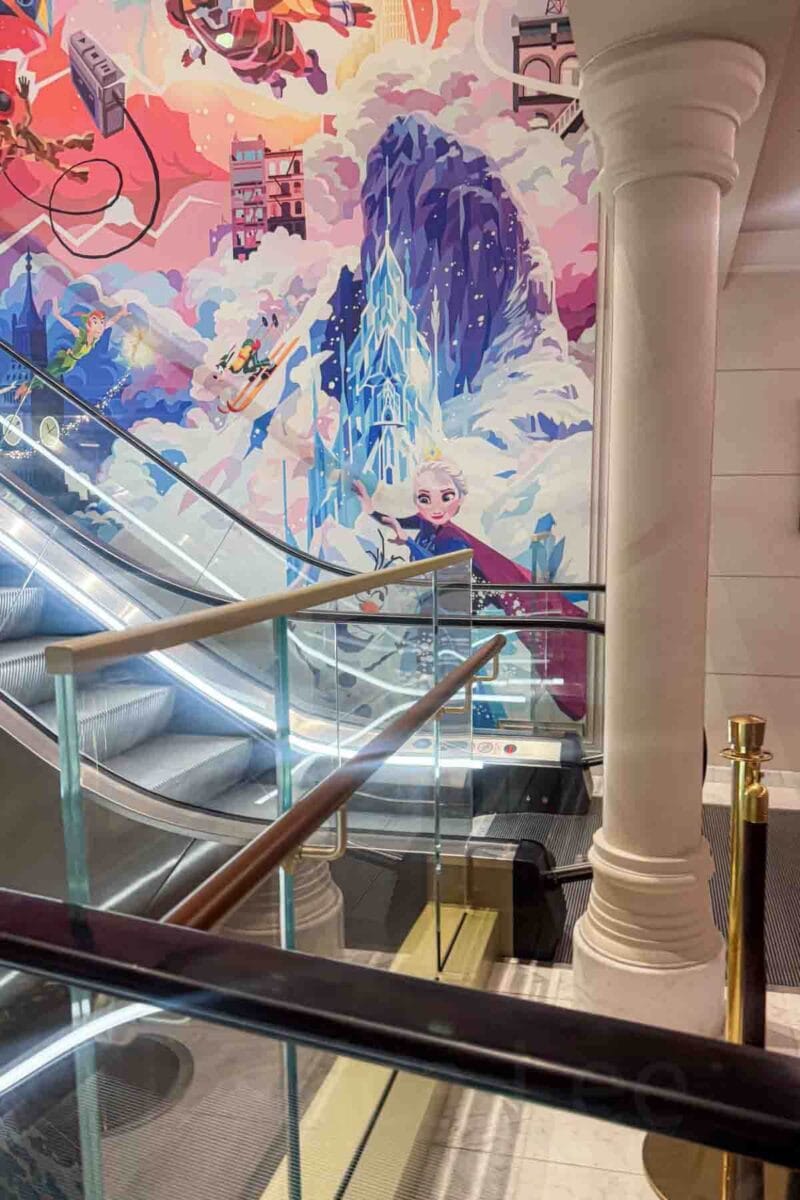 An escalator passes a large wall mural featuring Elsa and a snowy castle with colorful animated scenes above with a partial peek at an Ironman feature. This space highlights themed interior design on a Disney cruise ship.