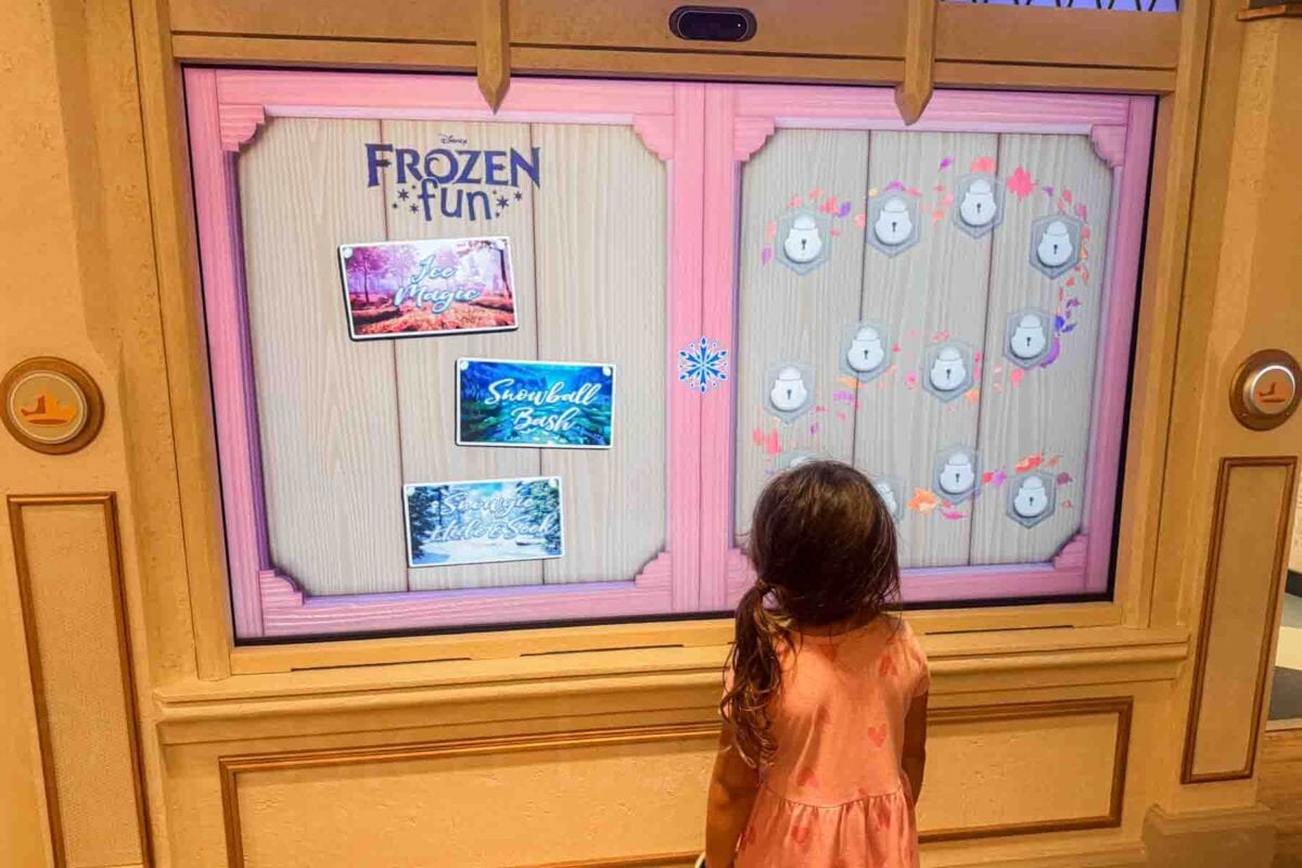 A young girl stands in front of a screen labeled "FROZEN FUN" with options including "Ice Magic", "Snowball Bash", and "Snowflake Stack". This interactive activity highlights kids club entertainment on a Disney cruise.