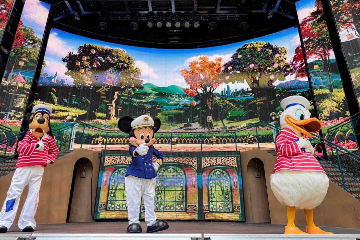Goofy, Mickey, and Donald stand on an outdoor stage with a vibrant garden themed digital backdrop. This performance highlights character entertainment on the Disney Adventure cruise.