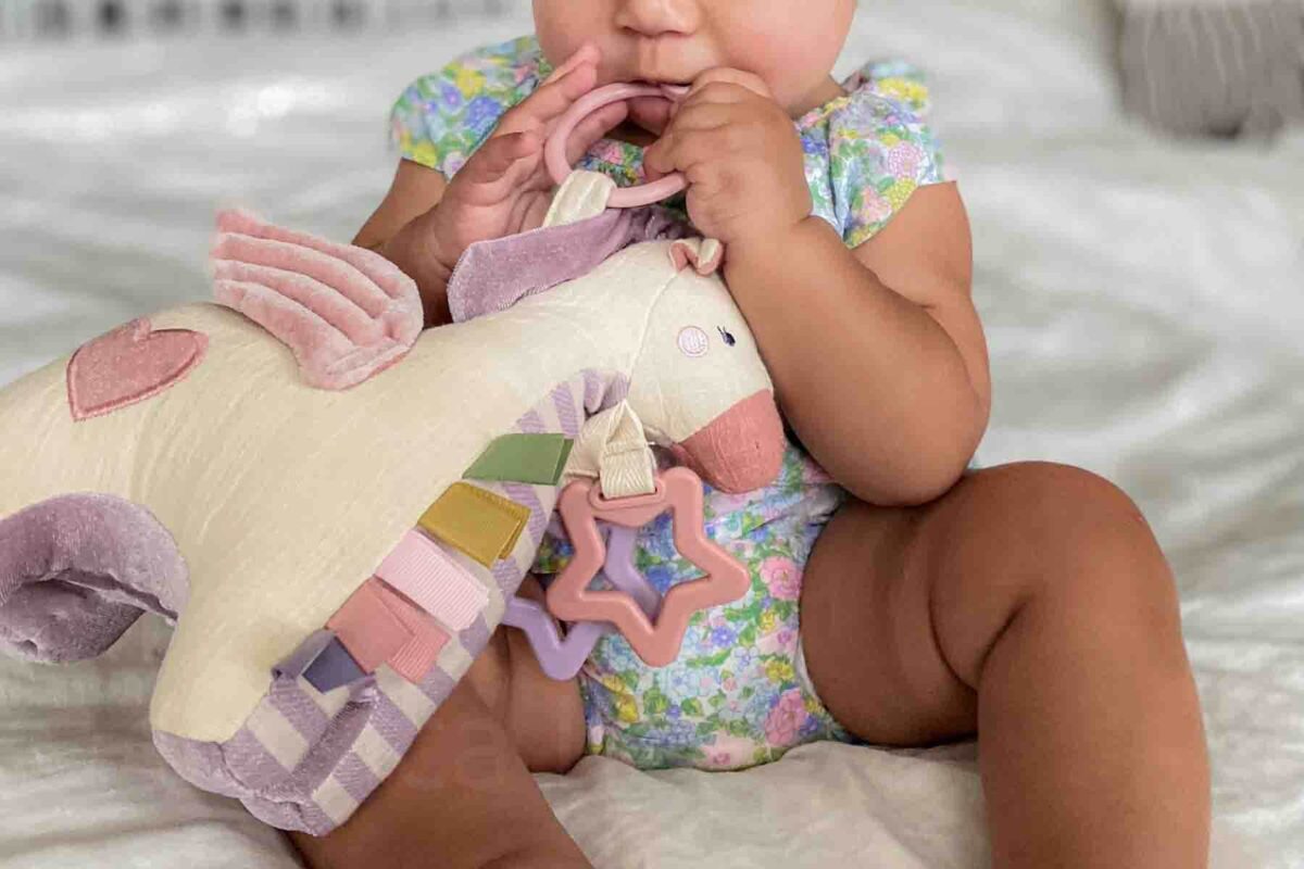 Baby holding and chewing on a teething ring of an Itzy Ritzy soft unicorn sensory toy with attached rings and textured fabric, a comforting all-in-one option among simple baby travel toys.