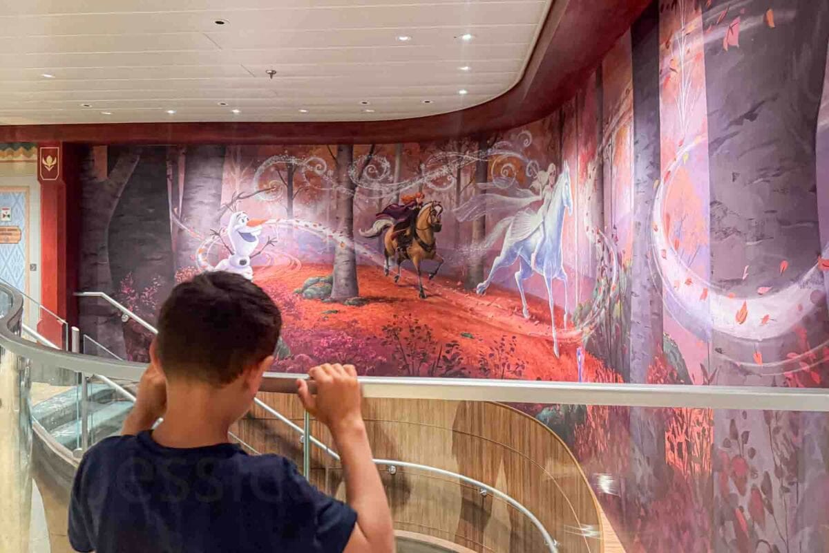 A child looks over a curved staircase railing at a large mural featuring Olaf, Anna, and Elsa in a forest scene from Frozen II. This artwork highlights immersive Frozen themed spaces on the Disney Adventure cruise.