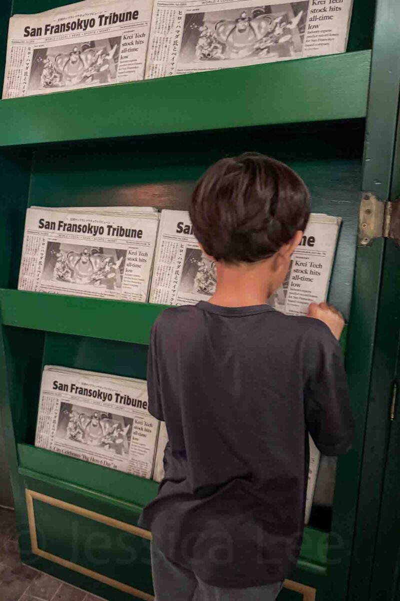 A child stands in front of shelves filled with faux newspapers labeled "San Fransokyo Tribune" with headlines reading "Krei Tech stock hits all time low". This interactive display highlights Big Hero 6 themed areas on the Disney Adventure cruise.