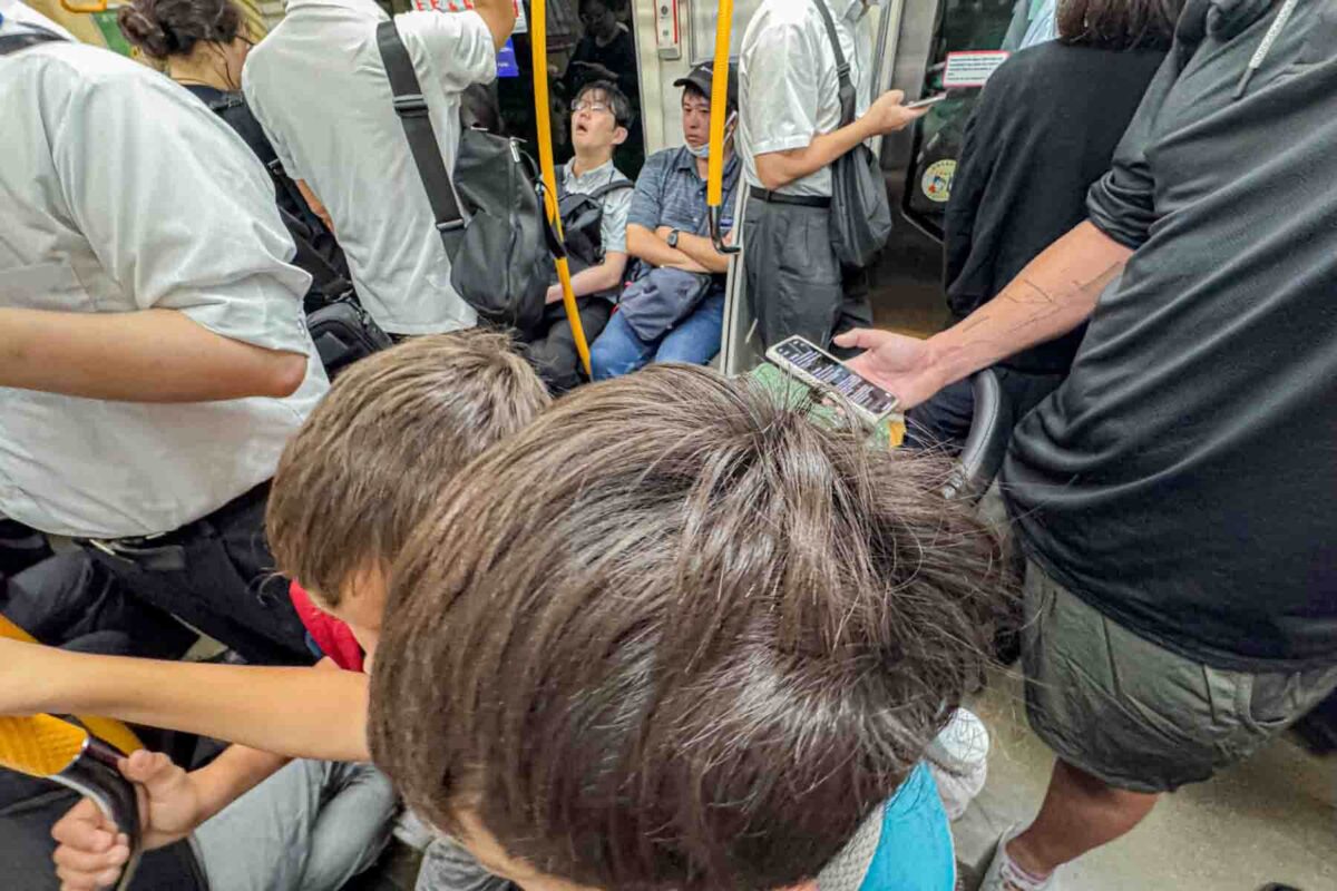 Two children stand surrounded by adults inside a crowded Tokyo subway car, holding onto poles during a busy rush hour commute. This scene reflects the reality of Tokyo public transportation with kids.