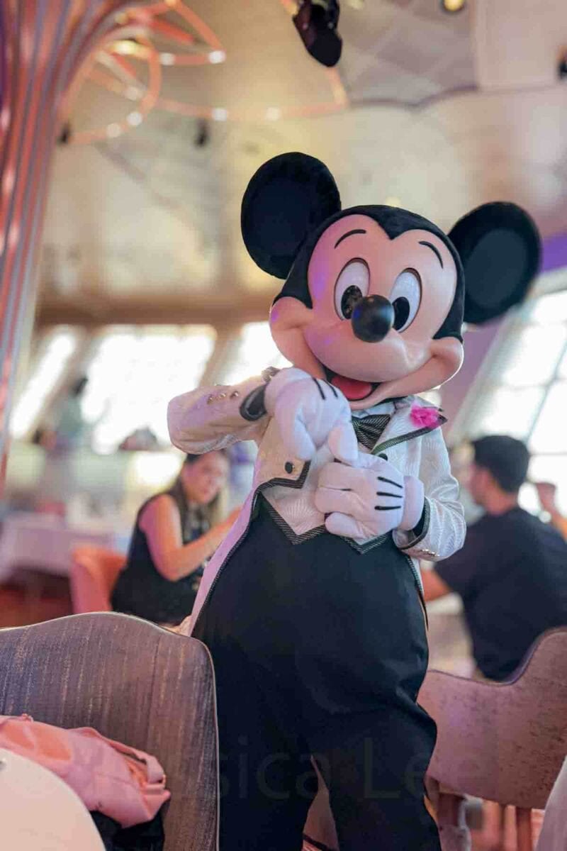 Mickey Mouse dressed in a white jacket and black bow tie poses and gestures while visiting guests in a dining room at the Hollywood Spotlight Club onboard the Disney Adventure. This character interaction highlights onboard experiences on a Disney cruise.