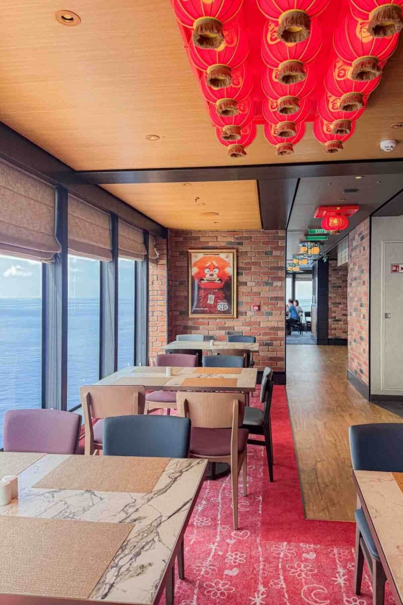 A restaurant dining area with red lanterns hanging from the ceiling and a "Turning Red" poster on a brick wall overlooks the ocean. This themed space highlights Pixar inspired dining on the Disney Adventure cruise.