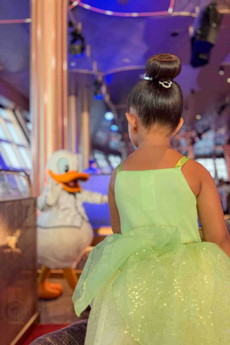 A young child in a green Tiana dress watches Donald Duck walking through a dining room during a character meal at the Hollywood Spotlight Club. This moment highlights family friendly dining entertainment on a Disney cruise.