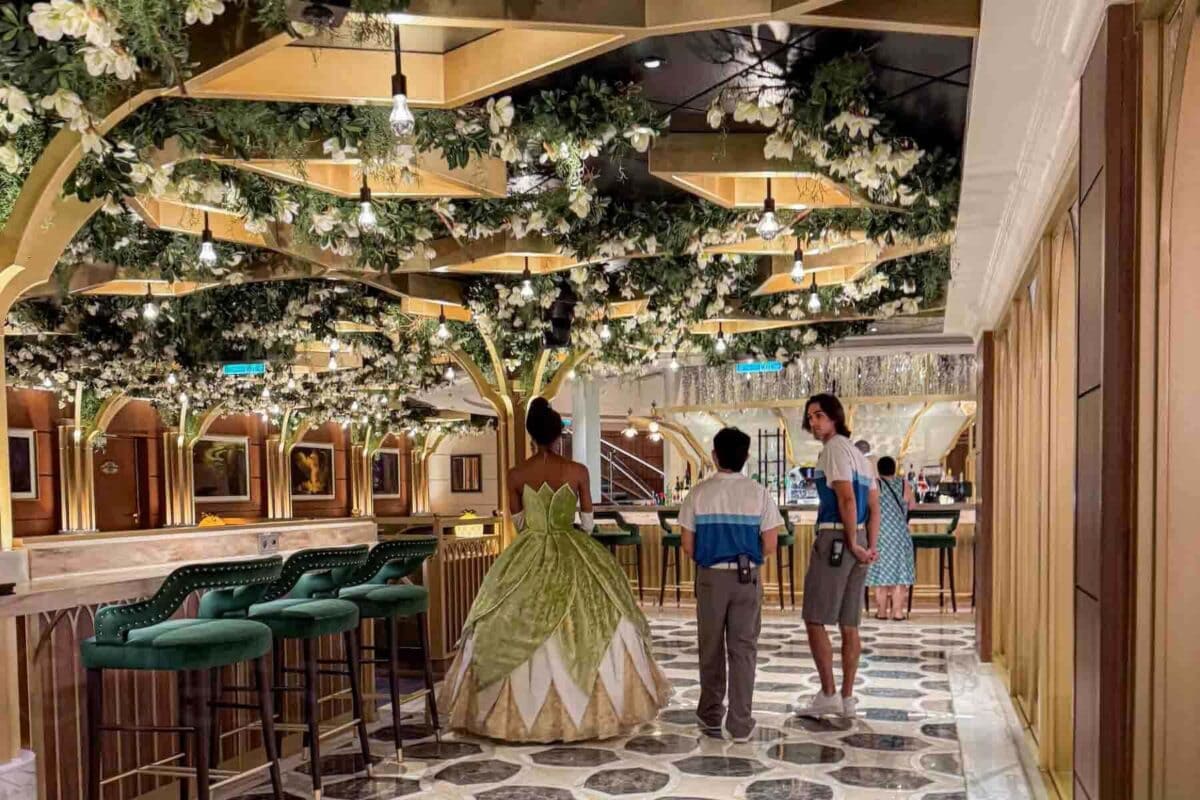 Princess Tiana walks was cast members standing near a bar in a lounge decorated with greenery, Magnolia flowers, and tree like columns overhead. This elegant bayou themed space highlights immersive dining and lounge design on the Disney Adventure cruise.