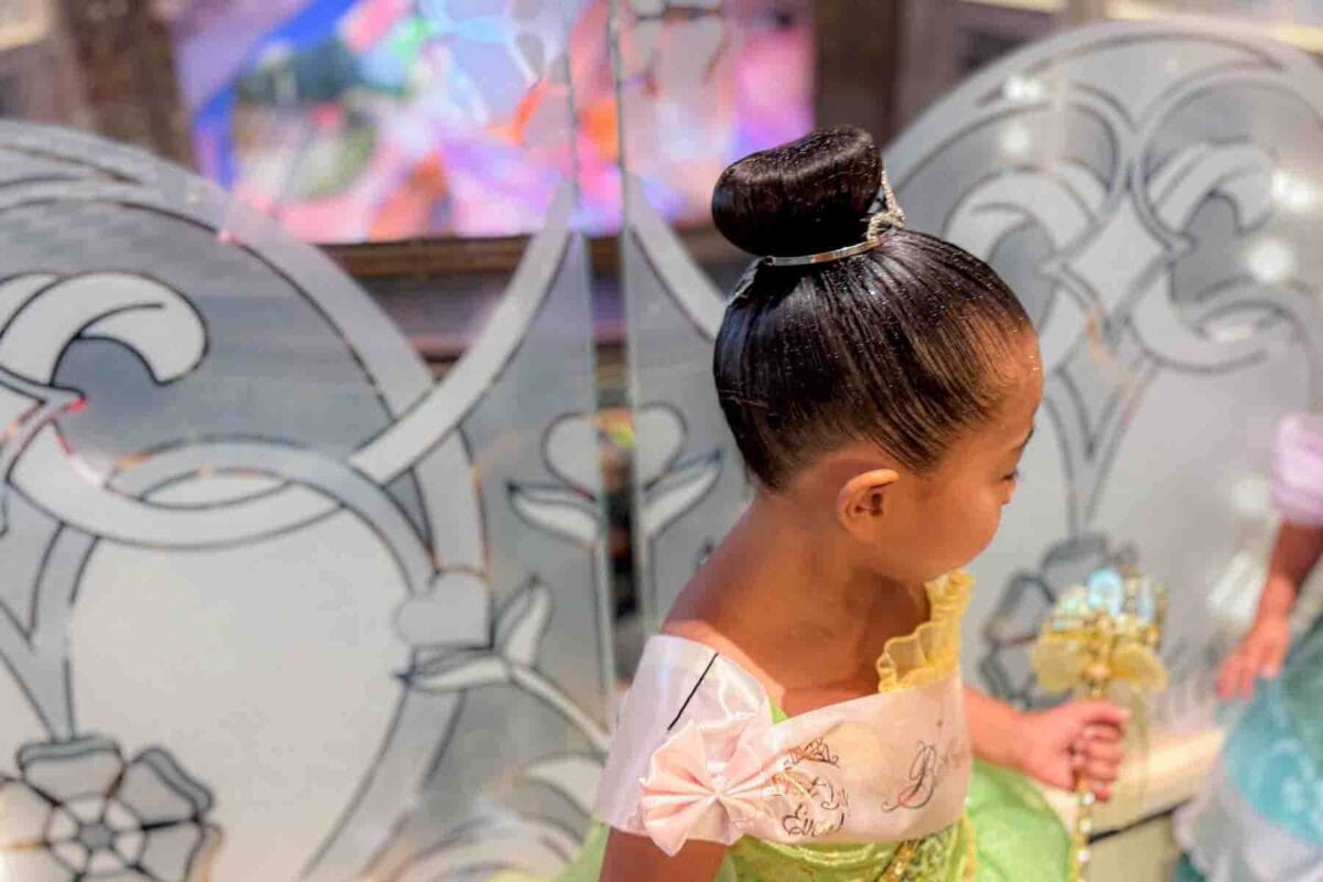 A young child dressed as Princess Tiana with a tiara and wand looks to the side while wearing a green gown. This moment highlights princess dress up experiences at Bibbidi Bobbidi Boutique on a Disney cruise.
