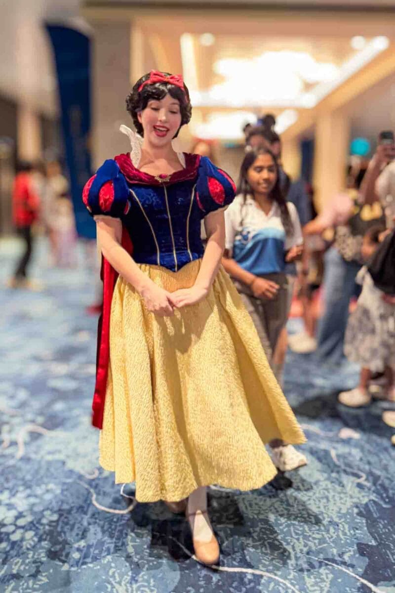 Snow White walks through a cruise ship corridor on a blue patterned carpet while guests watch nearby. This character appearance highlights onboard entertainment and meet and greet experiences.