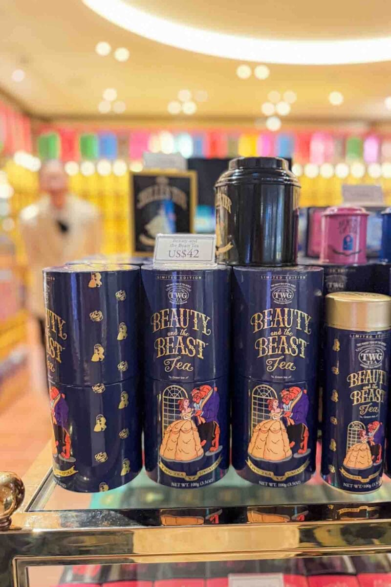 Stacked tins labeled "Beauty and the Beast Tea" with illustrated characters are displayed on a shop shelf with a sign reading "US$42". This merchandise highlights themed souvenirs available on the Disney Adventure cruise.