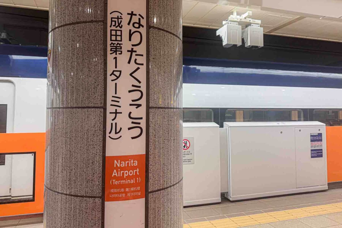 A pillar sign on a Tokyo train platform reads "Narita Airport Terminal 1" in English and Japanese beside a stopped train. Clear signage like this makes getting around Tokyo with kids and navigating routes easier.
