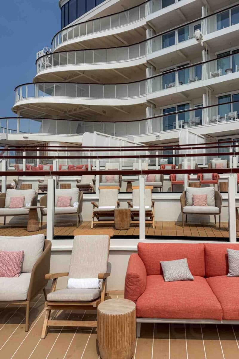 Rows of cushioned lounge chairs and sofas are arranged on an outdoor deck with glass railings and ship balconies above. This relaxing area highlights outdoor seating and views on a Disney Adventure cruise.