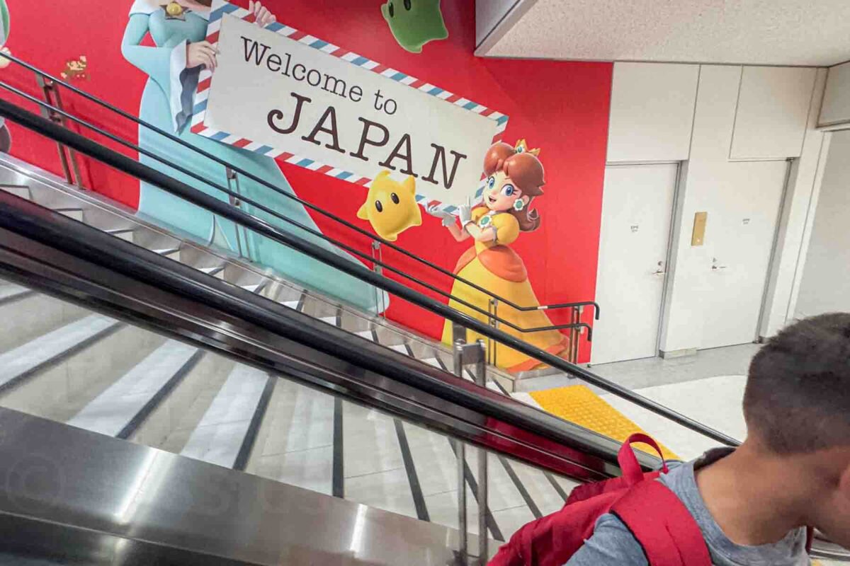 A "Welcome to Japan" sign on a wall mural held by Princess Peach and other Mario characters running along the stairwell and escalator in an airport train station near Tokyo with a small clip of a child wearing a backpack riding the escalator down.