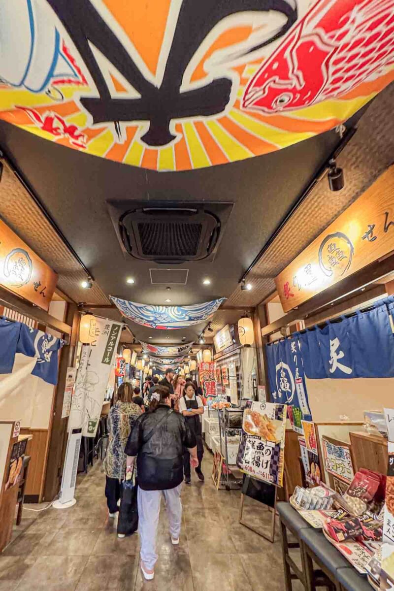 Narrow indoor food market hall next to Toyosu Market with Japanese lanterns, colorful ceiling banners, and small restaurant stalls lining both sides while visitors walk through the corridor. A lively place for families visiting Tokyo with kids to try local Japanese food in a casual setting.