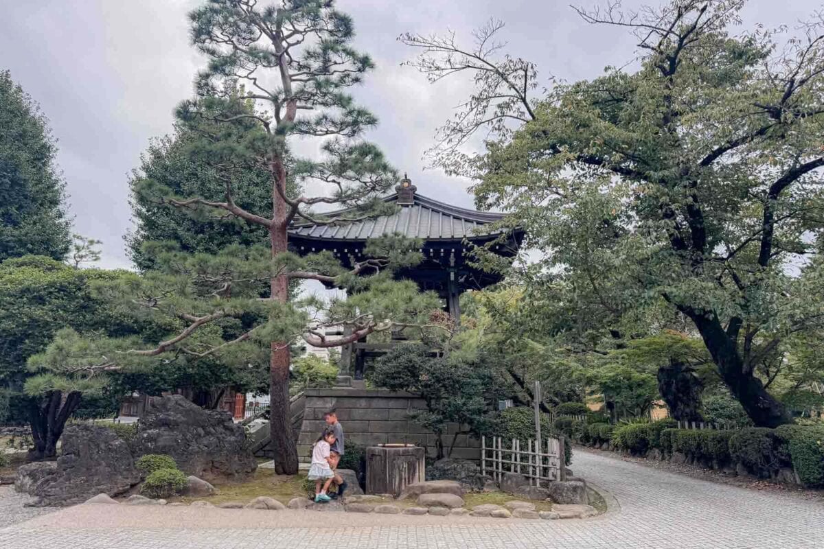 Two children standing beside a stone structure and pine tree with a temple roof peeking through inside the peaceful Gotokuji Temple grounds in Tokyo. Quiet temple gardens like this can offer a calm break for families exploring the city with young kids.