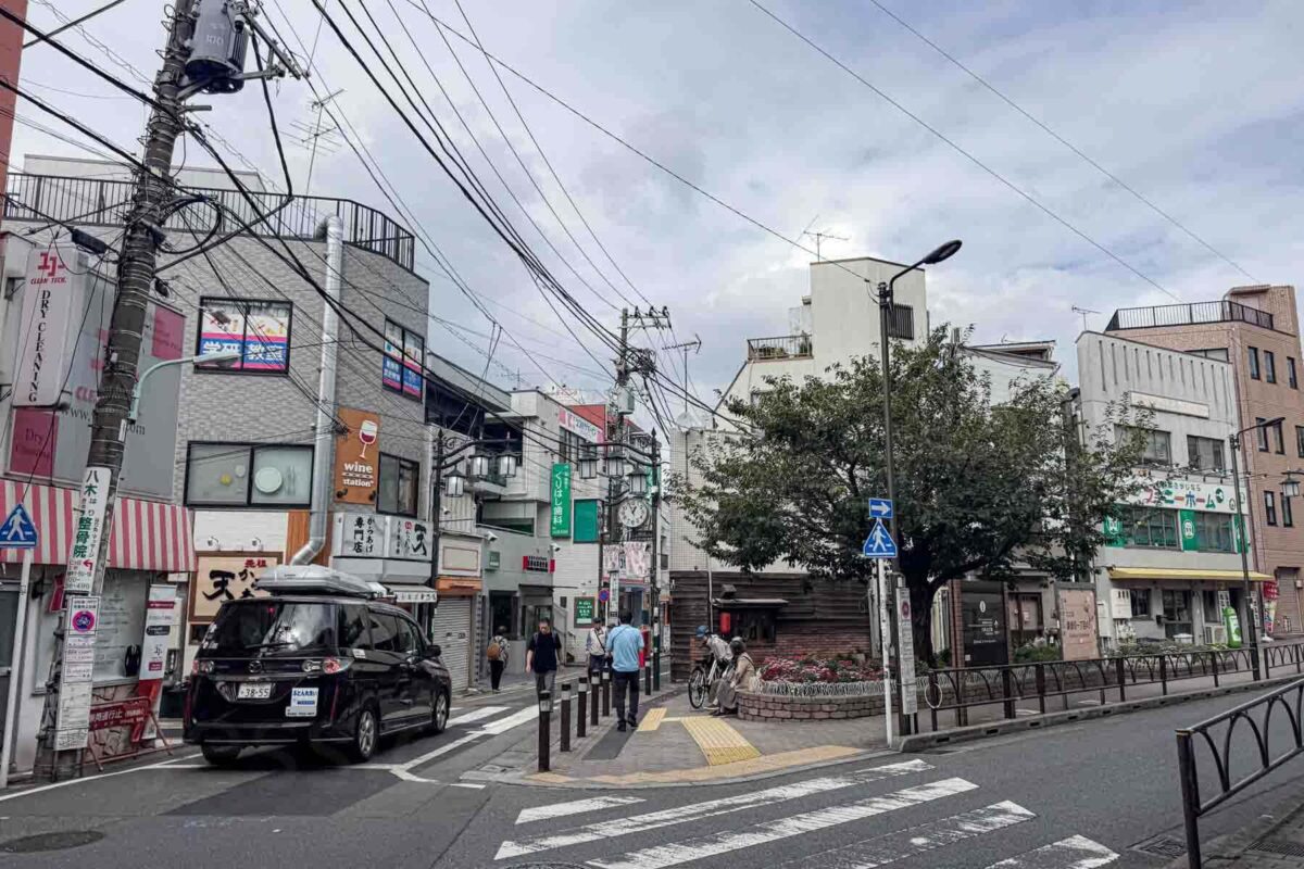 Residential Tokyo neighborhood street in Gotokuji with small shops, utility lines, bicycles, and pedestrians crossing near a quiet intersection. This everyday city scene shows what to expect in Tokyo with kids outside the main tourist areas.