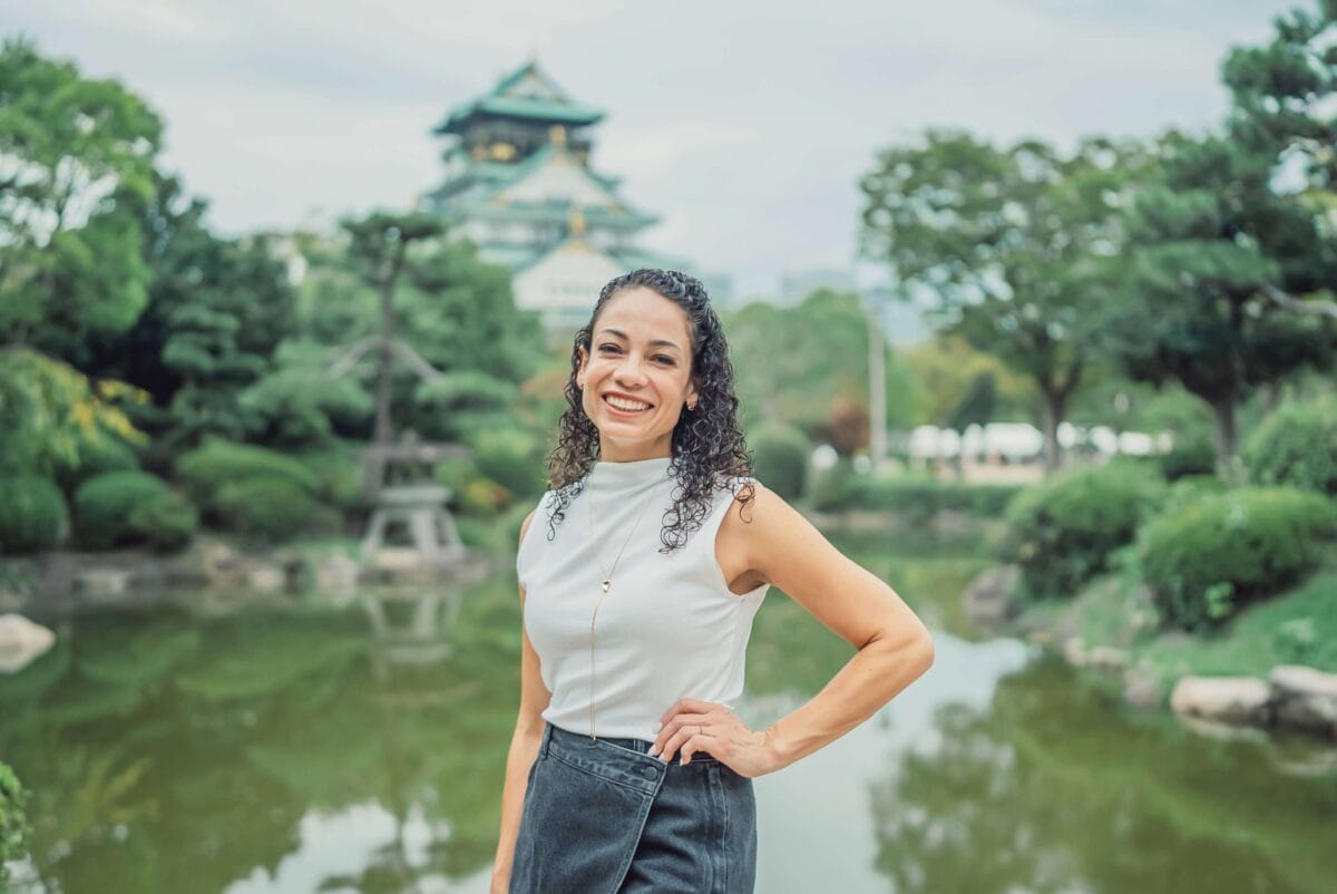 Woman wearing a white top with dark curly hair smiling with hand on hip posing in front of Osaka Castle in Japan
