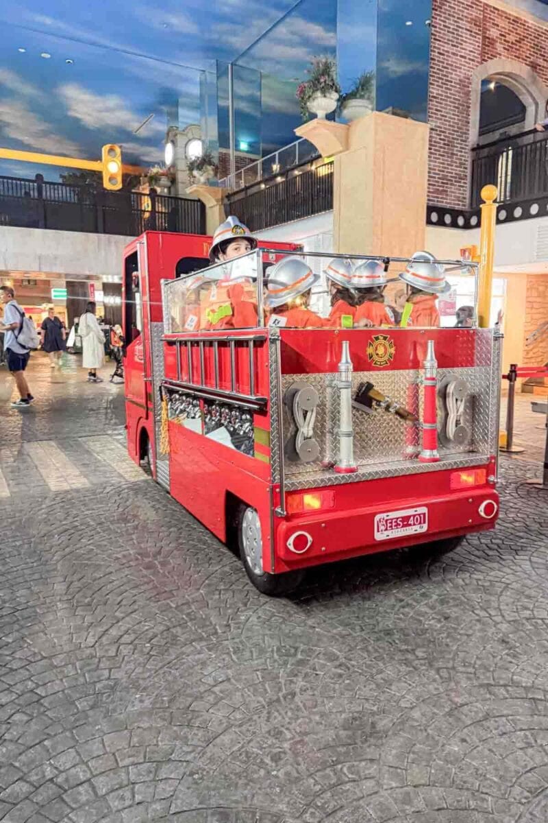 Children dressed as firefighters riding in a small red fire truck inside KidZania Tokyo during an interactive role play activity. Kid focused attractions like this are a highlight in a Tokyo with kids travel guide.