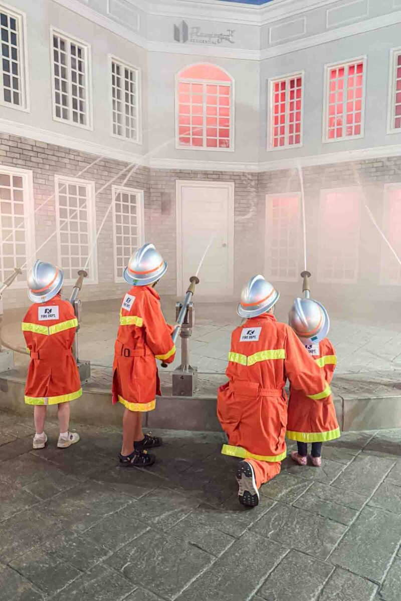 Children wearing firefighter uniforms and helmets spraying water hoses at a pretend building during a KidZania Tokyo emergency training activity. Interactive experiences like this make KidZania a memorable stop for families in Tokyo.