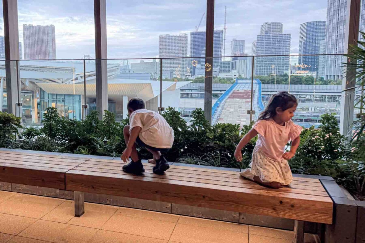 Two children playing on a wooden bench inside a modern Tokyo shopping mall with large windows overlooking the city skyline and waterfront. Indoor rest areas like this are helpful when getting around Tokyo with kids during busy sightseeing days.