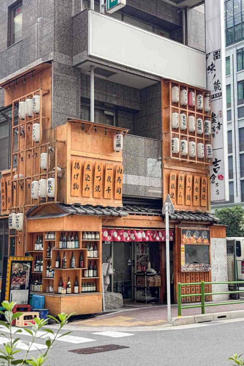 Traditional style Tokyo izakaya restaurant exterior with wooden signs, hanging lanterns, and shelves of sake bottles along the street corner. Small local restaurants like this add to the atmosphere of exploring all sides of Tokyo.