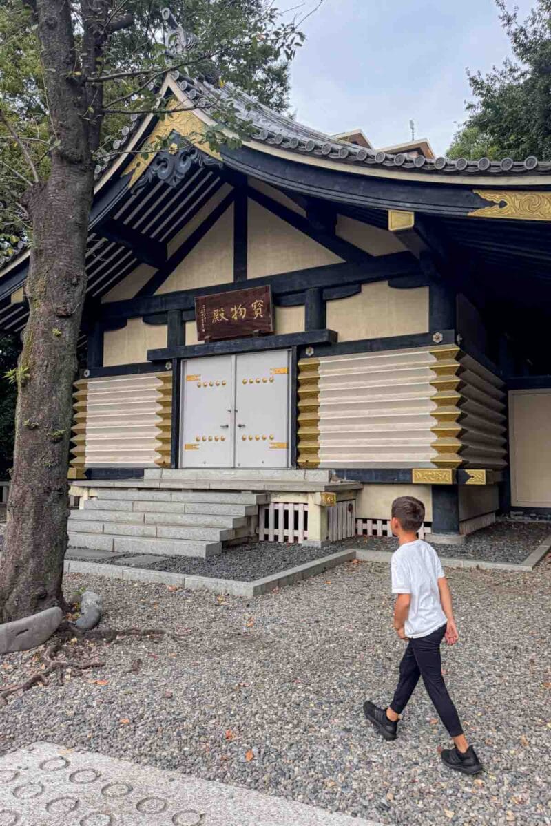 Child walking toward a traditional Japanese temple building with curved roof details and gold accents surrounded by trees in Tokyo. Historic temples like this offer a quieter cultural experience for families traveling through Tokyo.