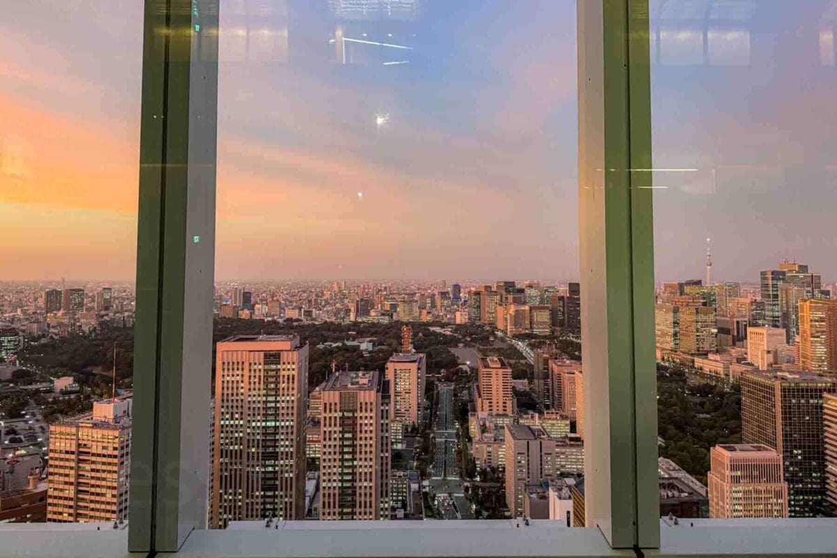 Wide skyline view of Tokyo at dusk seen through large observation windows with city buildings stretching into the distance under a pink and orange sky. Observation decks like this are a relaxing way for families to experience Tokyo from above the city.