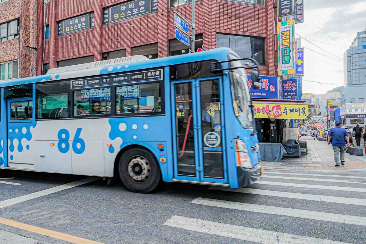 A blue and white Busan city bus marked "86" turns through an intersection surrounded by Korean storefronts. Buses help fill transportation gaps when the Metro does not reach a family destination directly.