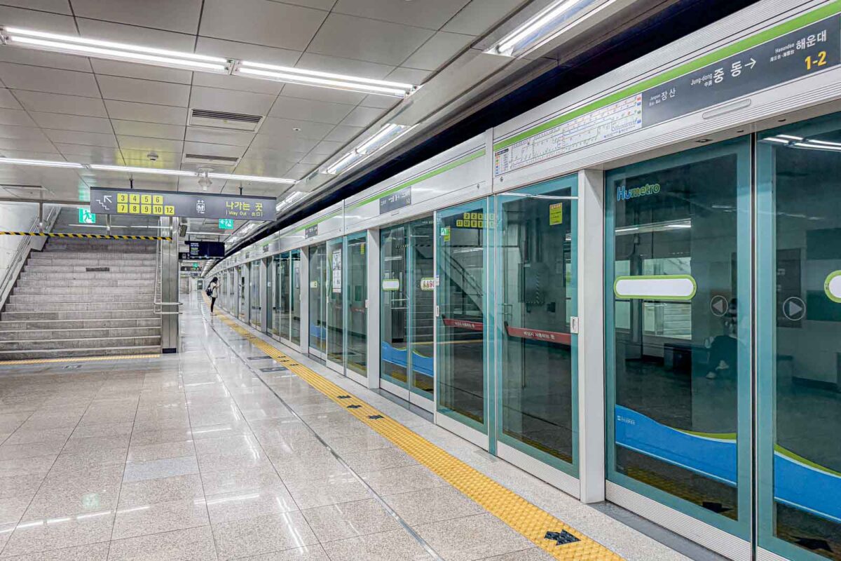 Glass platform screen doors separate passengers from the tracks at Jungdong Station on Busan Metro Line 2. Overhead signs show the direction toward "Jung dong" and "Haeundae" and mark exits "1 2".