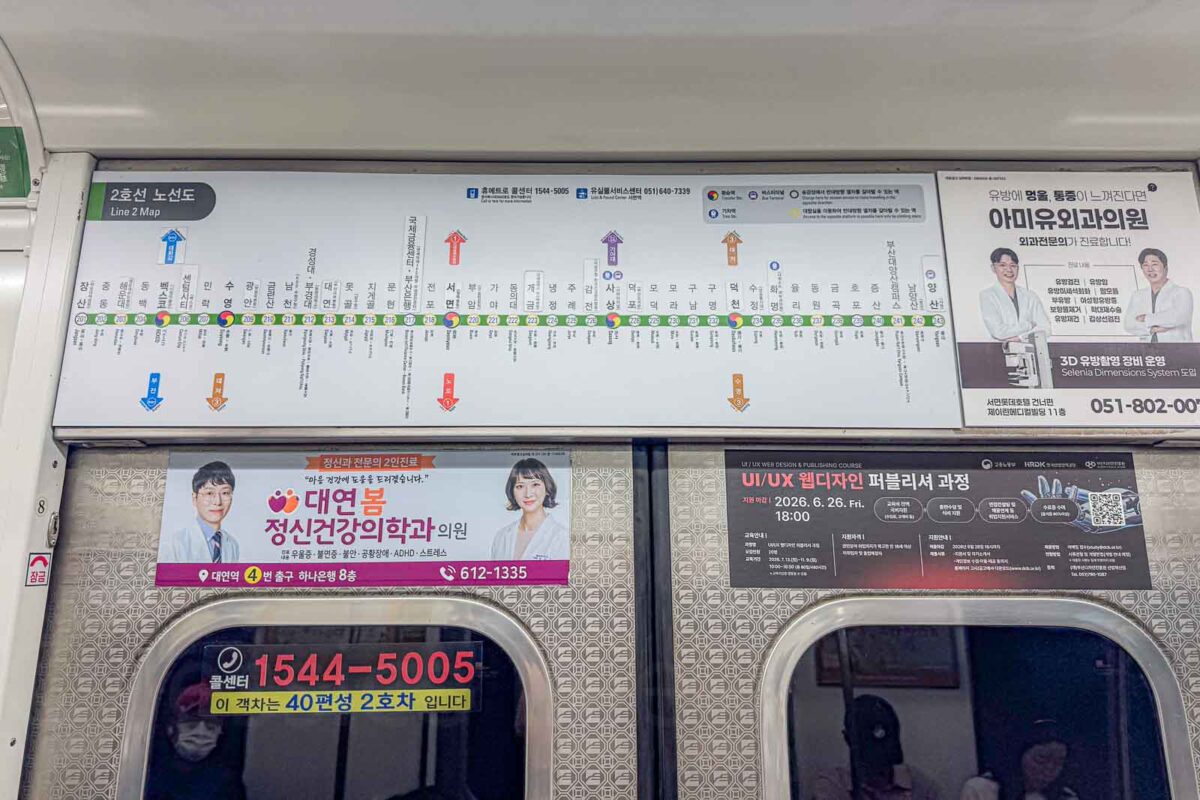 A long green route diagram above the doors of a Busan Metro train maps the stations and transfers on "Line 2 Map". The diagram helps passengers follow their progress and identify connections while using Busan public transportation.