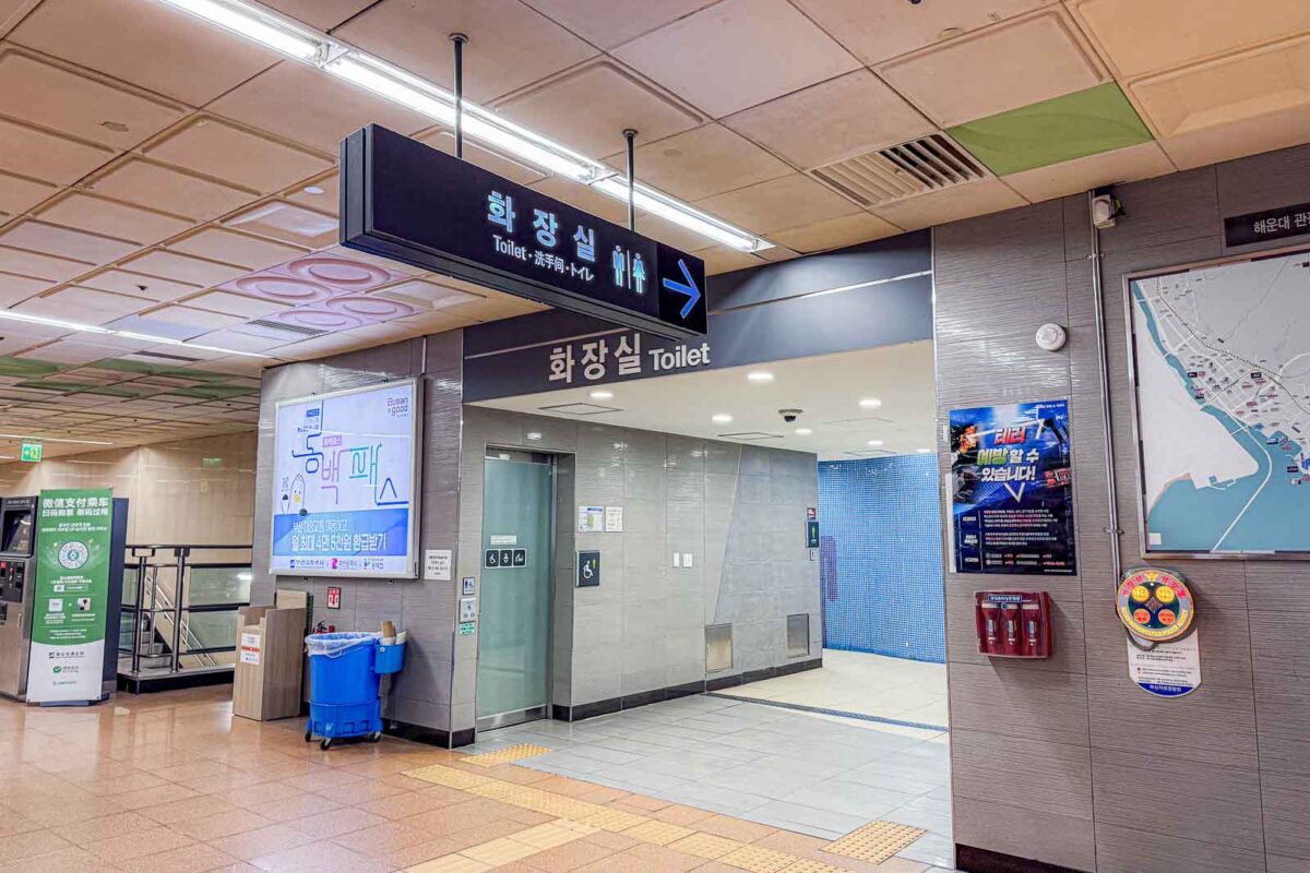 An overhead sign inside a Busan Metro station points toward public restrooms. The signs read "Toilet" in English beside male and female restroom symbols.