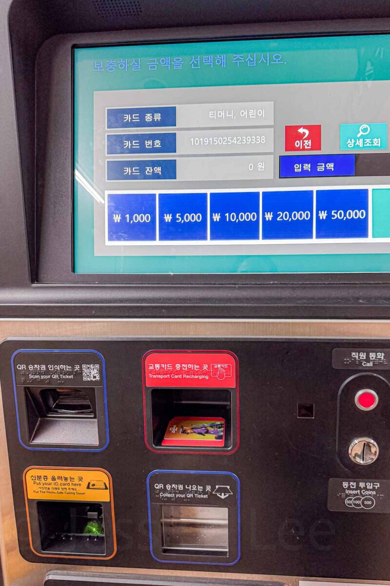 A Busan Metro machine displays options for adding money to a transportation card with buttons for "₩ 1.000". "₩ 5.000". "₩ 10.000". "₩ 20.000". "₩ 50.000". Labels on the machine include "Scan your QR Ticket". "Transport Card Recharging". "Put your IC card here". "Collect your QR Ticket". "Insert Coins". "Call".