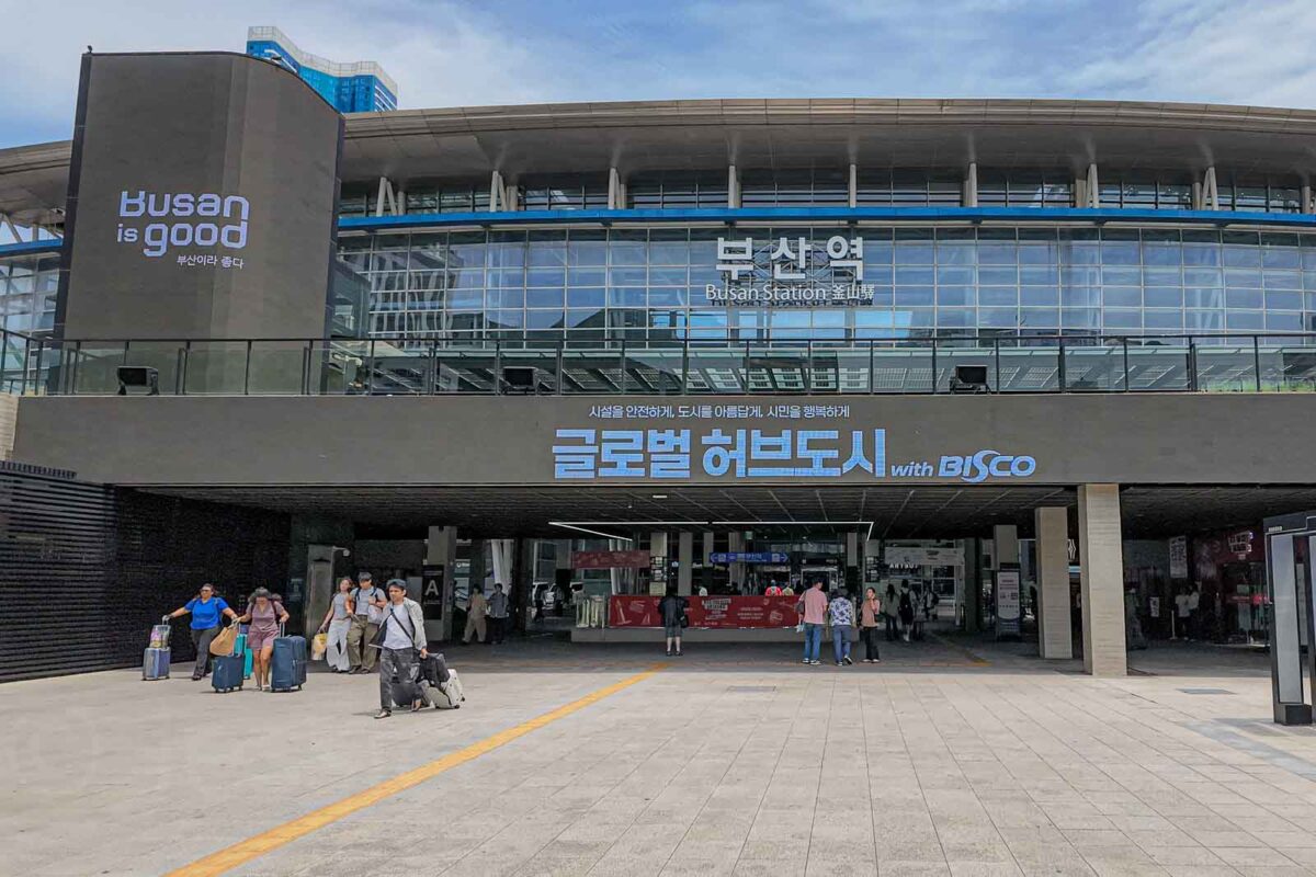 Travelers with rolling luggage gather outside the large glass facade of Busan Station. Signs read "busan is good". "부산역". "Busan Station". "글로벌 허브도시 with BISCO".