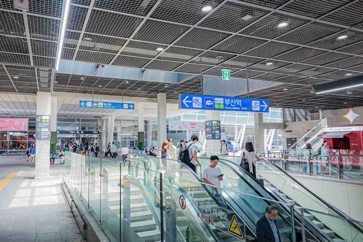 Travelers move between escalators inside the spacious main hall of Busan Station beneath blue directional signs. One large sign reads "부산역". "Busan Station". The open layout and escalators help families move between trains and local transportation with luggage.