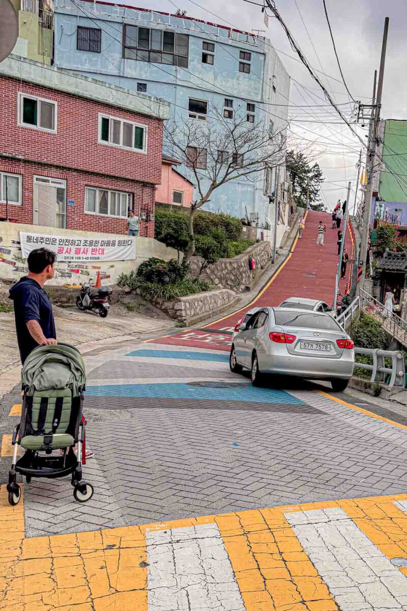 An adult pushes a stroller down a steep road at Gamcheon Culture Village while a car climbs the narrow hill ahead. The sharp slopes and shared road space show why this neighborhood is one of the more difficult places to explore in Busan with a stroller.