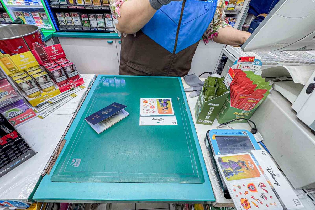 Two colorful Disney-themed T-money cards and passports sit on a convenience store counter while an employee registers them. The photo shows the process this family used to get registered child T money cards in Busan.