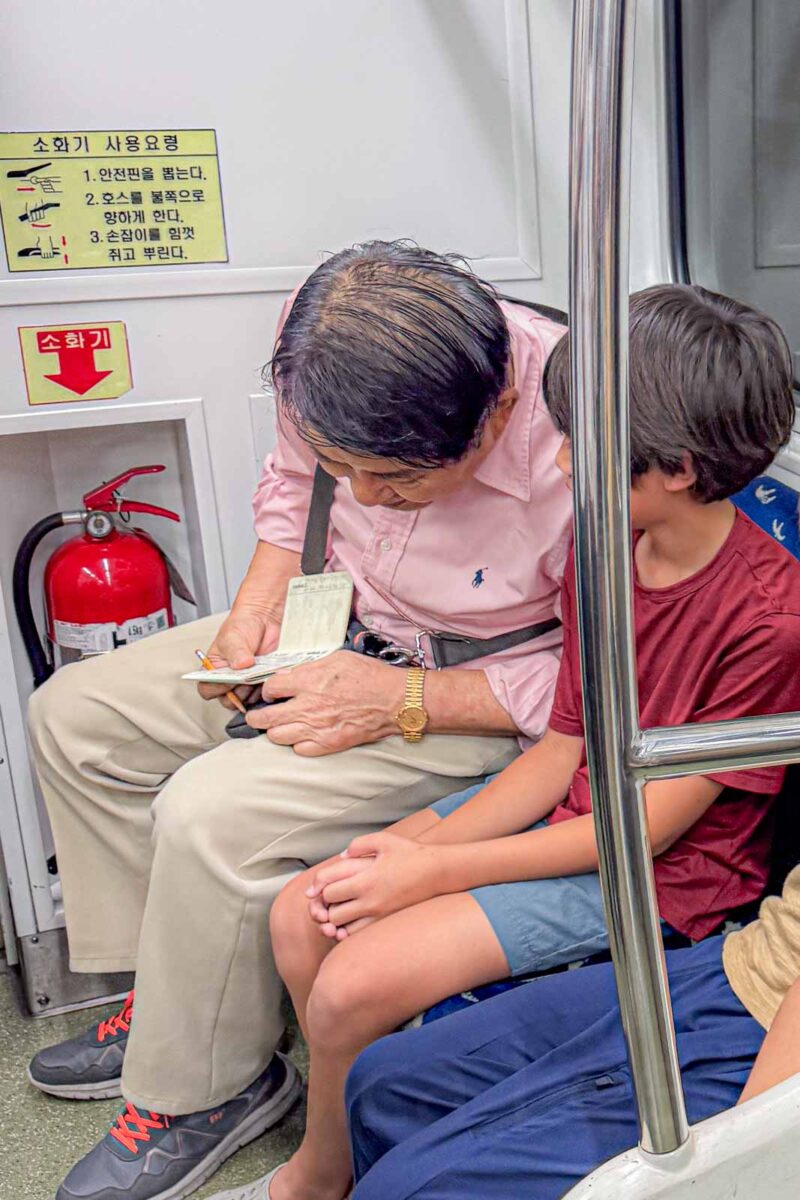 An older passenger sits closely beside a child on a Busan Metro train while looking through papers in his hands. The interaction reflects the friendly encounters this family experienced with other passengers while riding the Metro with kids.