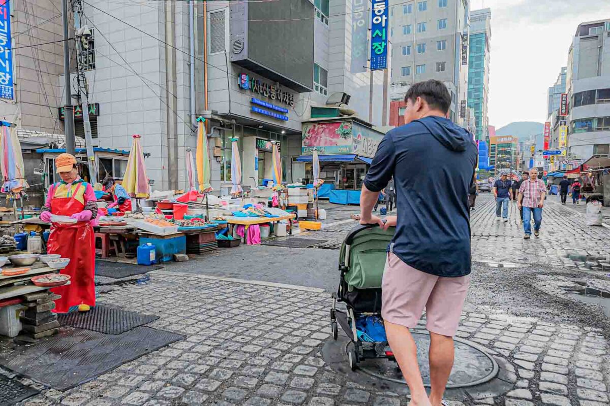 An adult pushes a green stroller over a wet uneven street beside seafood vendors outside Jagalchi Market. The busy market surroundings show how walking conditions can change from the wide sidewalks found in newer parts of Busan.
