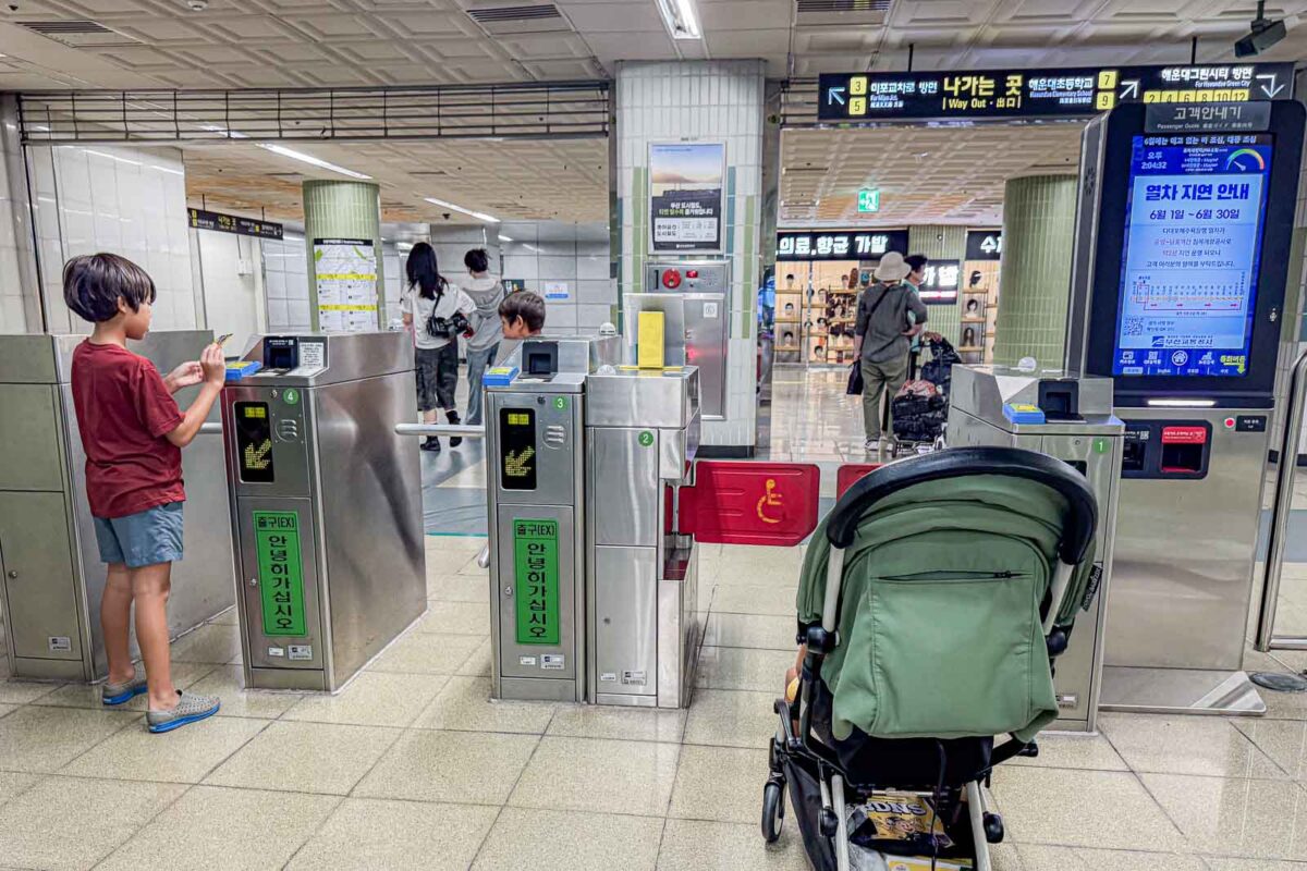 A child holds a transportation card beside Busan Metro fare gates while a green stroller waits in front of the wider accessible gate. The setup shows how families with strollers can use the Metro when getting around Busan with kids.