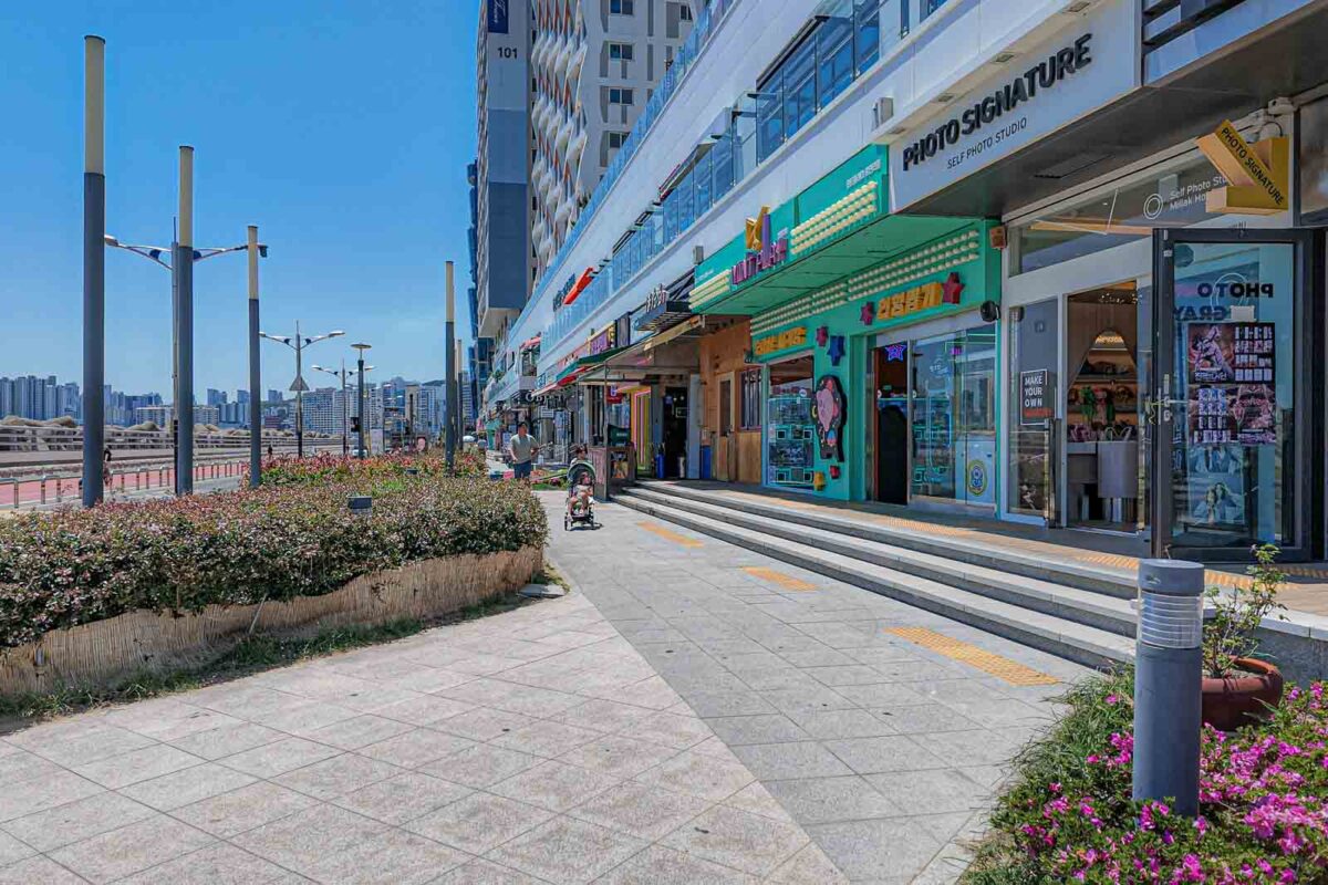 An adult pushes a stroller along a wide sunny sidewalk beside colorful storefronts near Gwangalli Beach. The smooth pavement and open walking space show one of the easier areas for navigating Busan with a stroller.