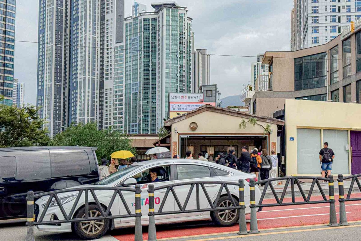 A taxi passes restaurants and low rise buildings beneath tall apartment towers in Haeundae. Taxis can provide convenient door to door transportation when weather. Distance. Or tired kids make walking less appealing.