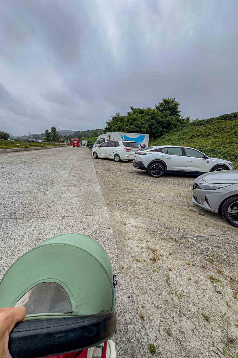 A hand pushes a compact green stroller along the edge of a rough gravel road beside parked cars. The uneven final stretch shows why families should check walking conditions as well as the bus or Metro portion of a Busan route.