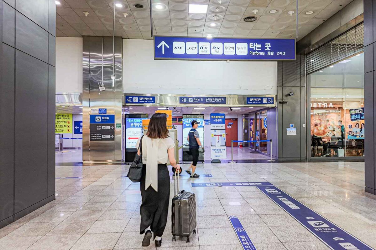 A traveler pulls a suitcase through Busan Station beneath a large blue sign pointing toward the railway platforms. The sign reads "KTX". "SRT". "ITX". "무궁화". "새마을". "타는 곳". "Platforms".