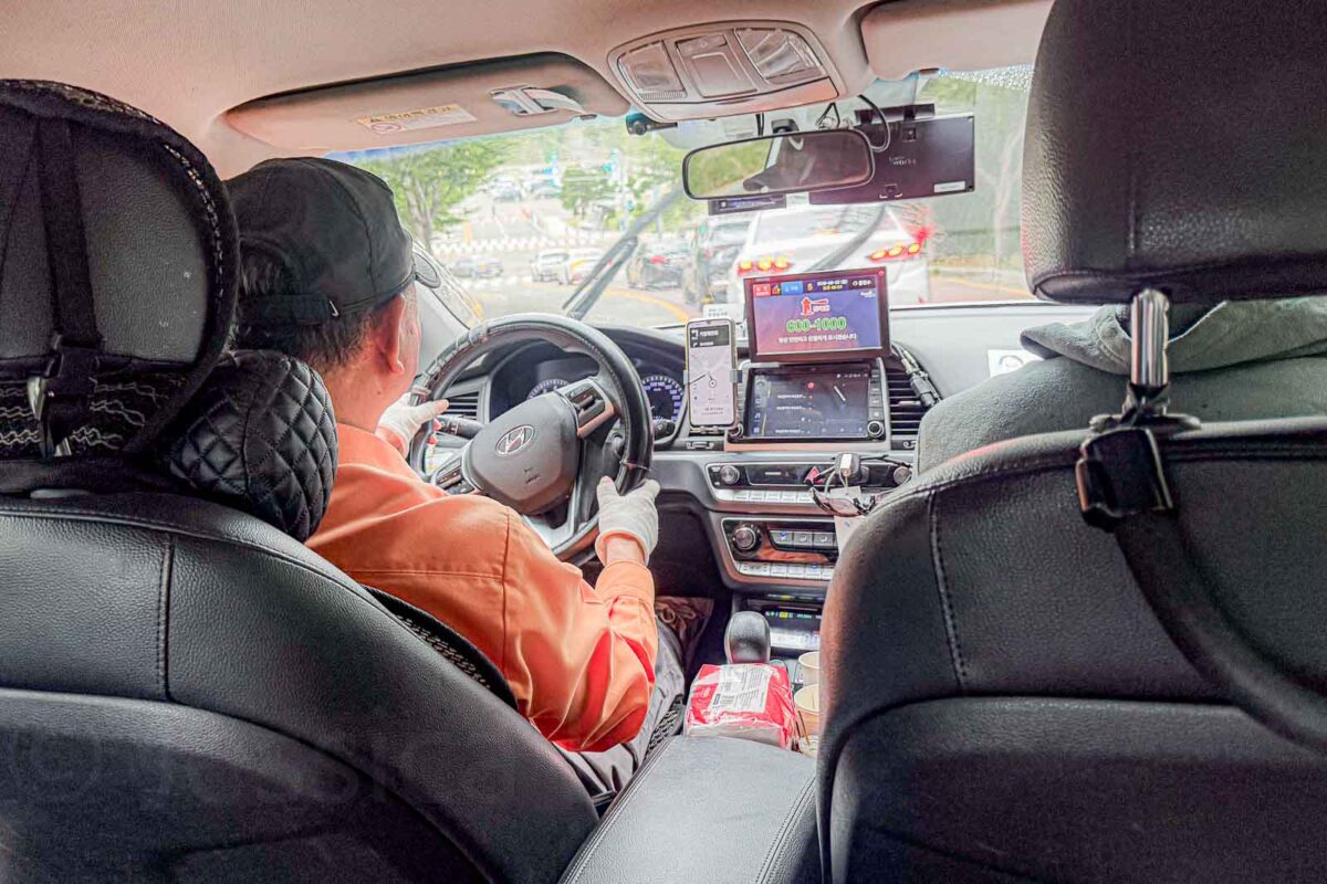 The interior of a Busan taxi shows the driver at the wheel with navigation screens and a phone mounted on the dashboard. The compact passenger space illustrates why families with several children may find standard Busan taxis a tight fit.