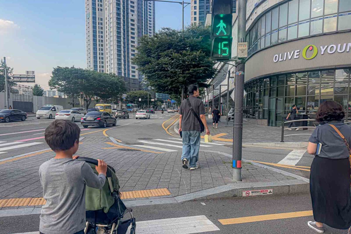  A child pushes a stroller across a Busan street beneath a green pedestrian signal displaying "15". The countdown helps families judge how much time remains to cross busy roads with young children.