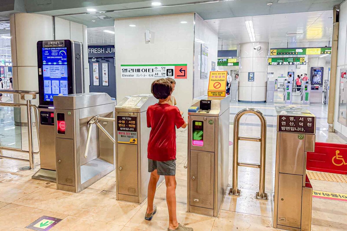 A child taps a transportation card at a Busan Metro fare gate before walking through. Nearby signs include "Elevator" and "Haeundae". Children who qualify for the free child fare still use their registered T money card at the gates.