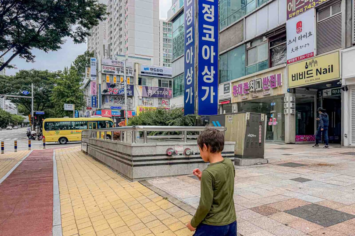 A child walks along yellow tactile paving on a broad sidewalk surrounded by shops and apartment buildings in Busan. A yellow bus is visible at the intersection ahead.