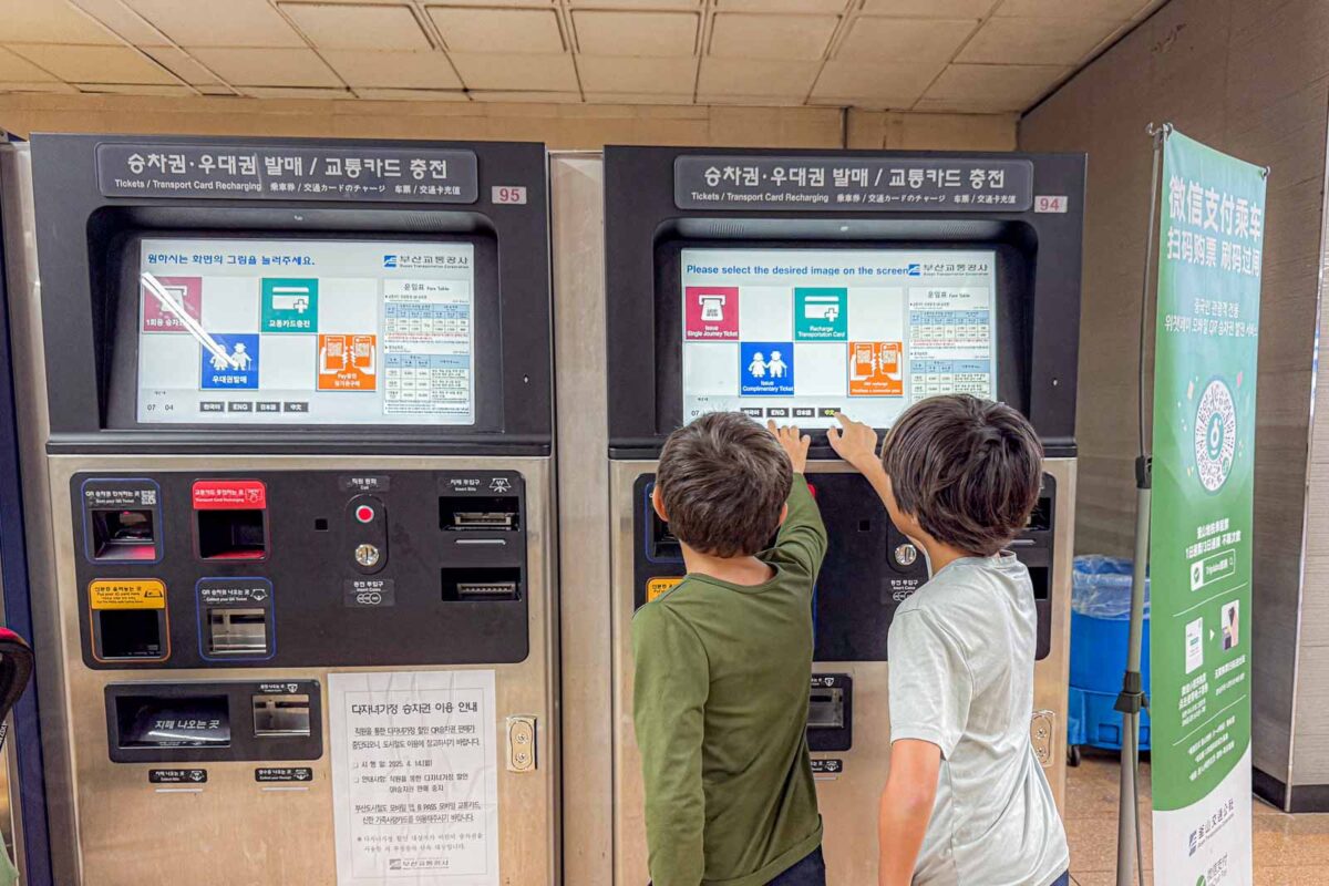 Two children use side by side ticket and transportation card machines inside a Busan Metro station. The machines display instructions in Korean and English including "Tickets Transport Card Recharging" and "Please select the desired image on the screen".