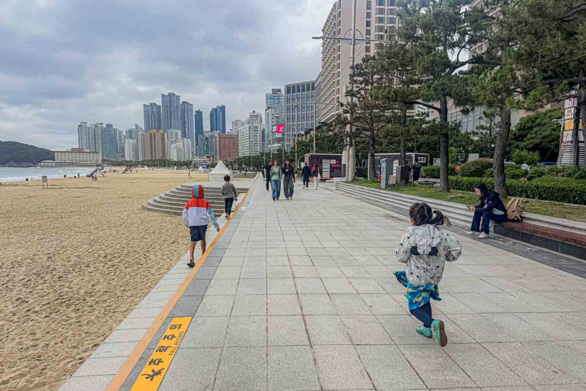 Children run along the broad paved promenade beside Haeundae Beach with high rise buildings lining the coast ahead. Flat pedestrian areas like this make Haeundae one of the easier Busan neighborhoods to explore on foot with kids.