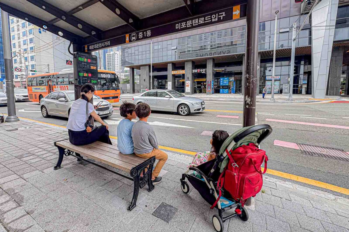 An adult and two children sit on a bench at a Haeundae bus stop while another child waits beside a loaded stroller. Orange city buses approach along the road in front of them.